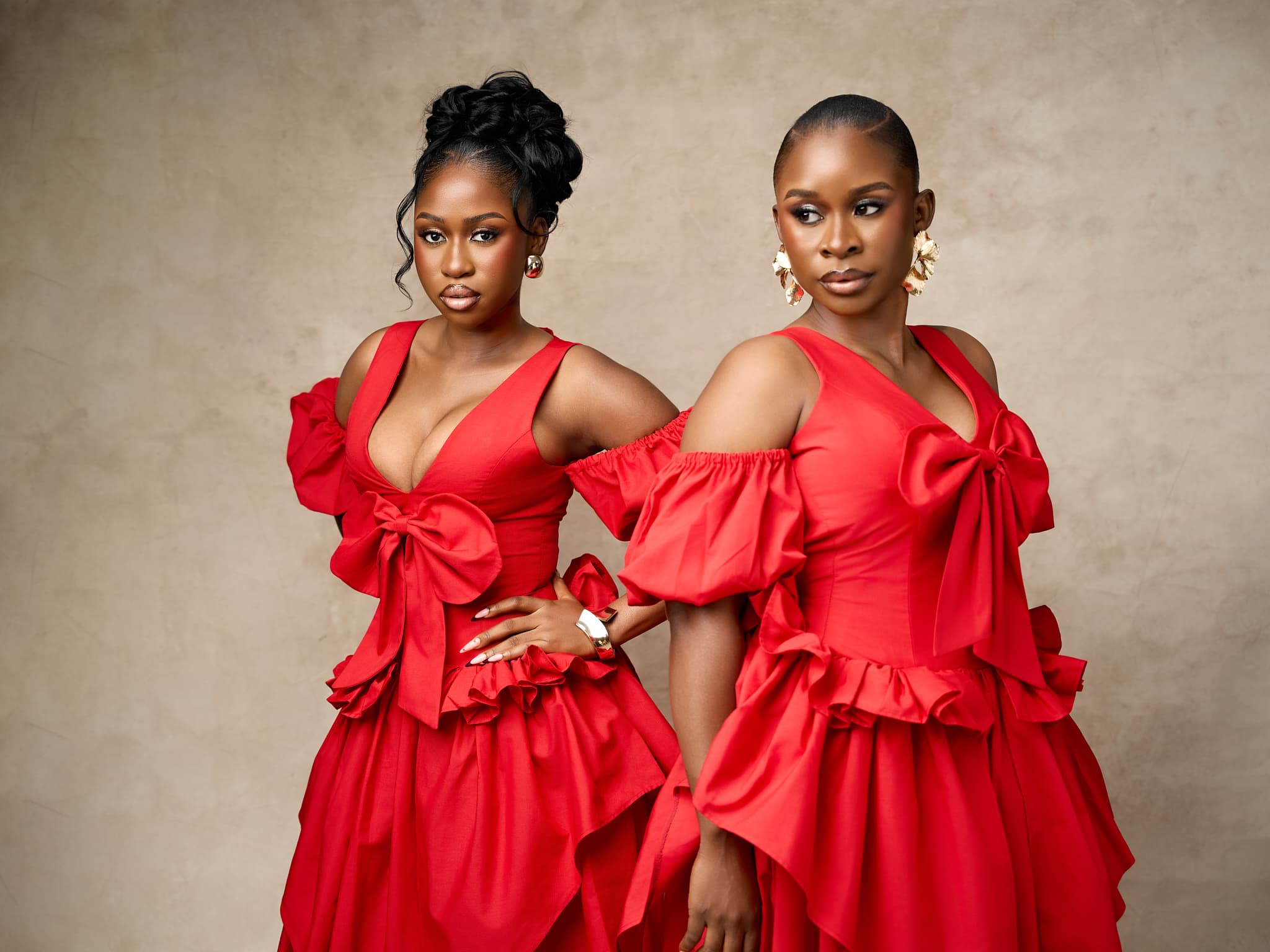 Two models in matching red dresses with oversized bow detailing and ruffled peplum skirts, against a warm stone backdrop.