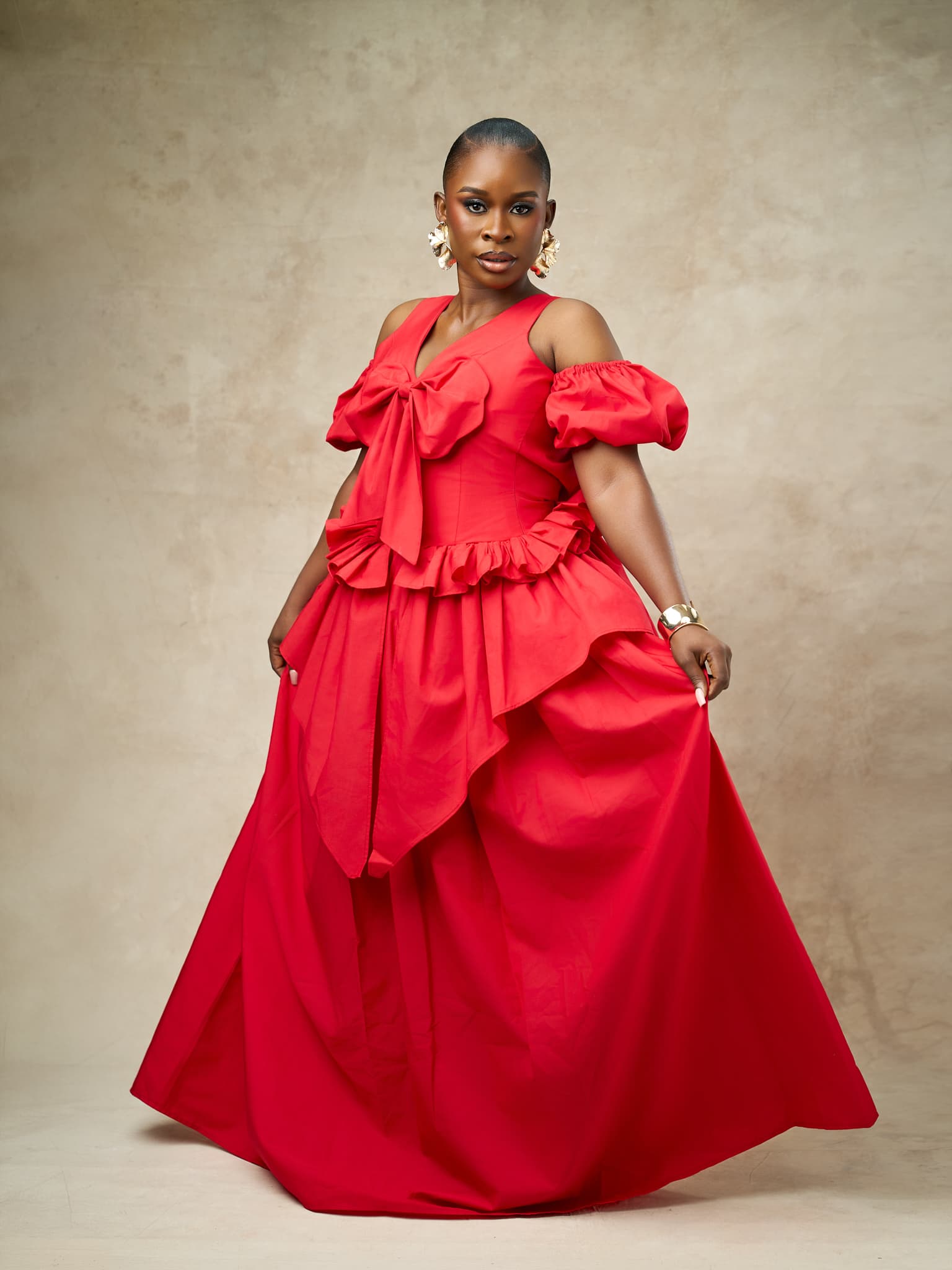 A model in a red gown with a bow at the bodice, off-shoulder puff sleeves and a full ruffled skirt, against a warm stone backdrop.