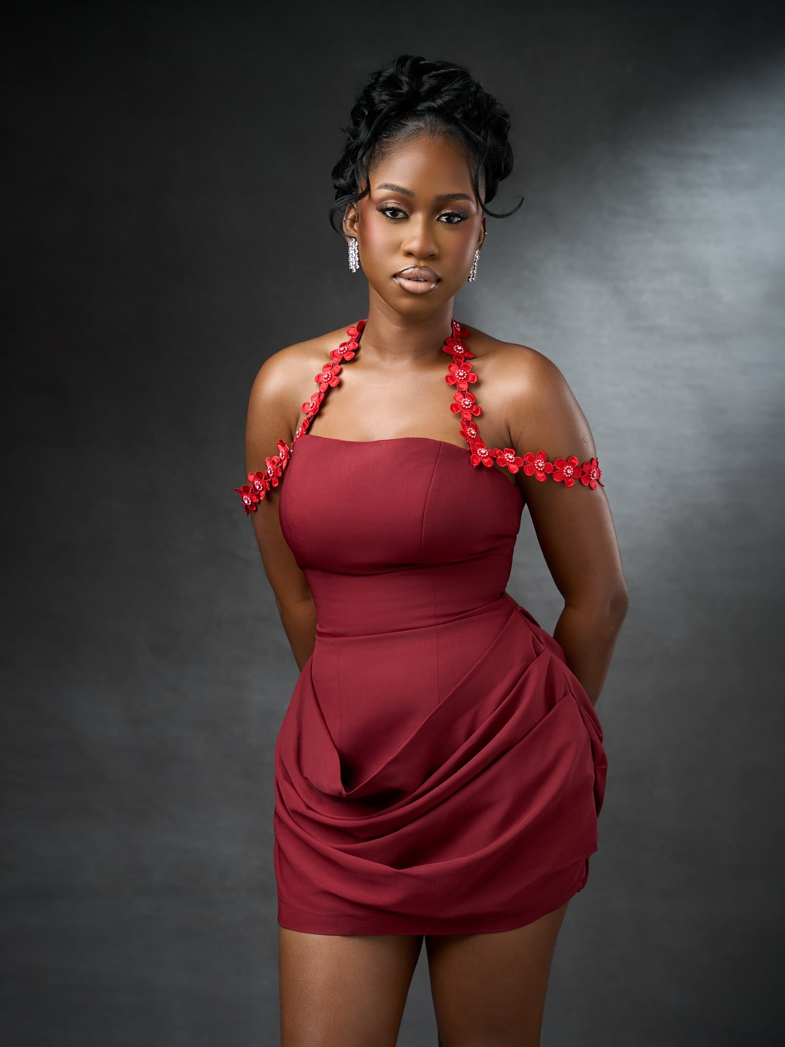 A model in a draped burgundy mini dress with a red floral off-shoulder strap, against a grey studio backdrop.