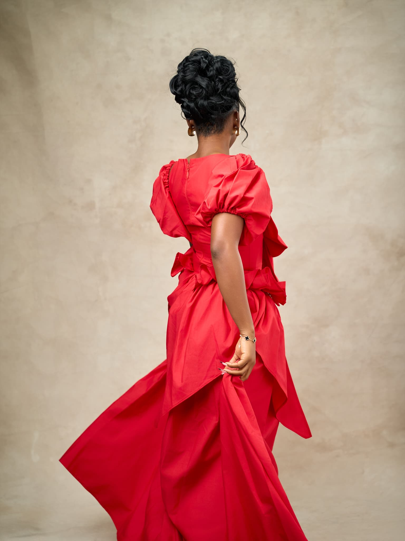 A model photographed from behind in a red gown with puff sleeves and a sweeping ruffled train, against a warm stone backdrop.
